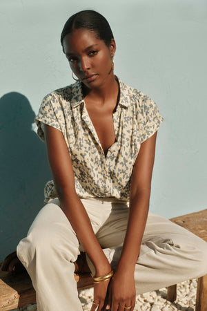 A black woman sitting on a bench wearing a Velvet by Graham & Spencer PAULETTE FLORAL BUTTON FRONT TOP with subtle stripe details.