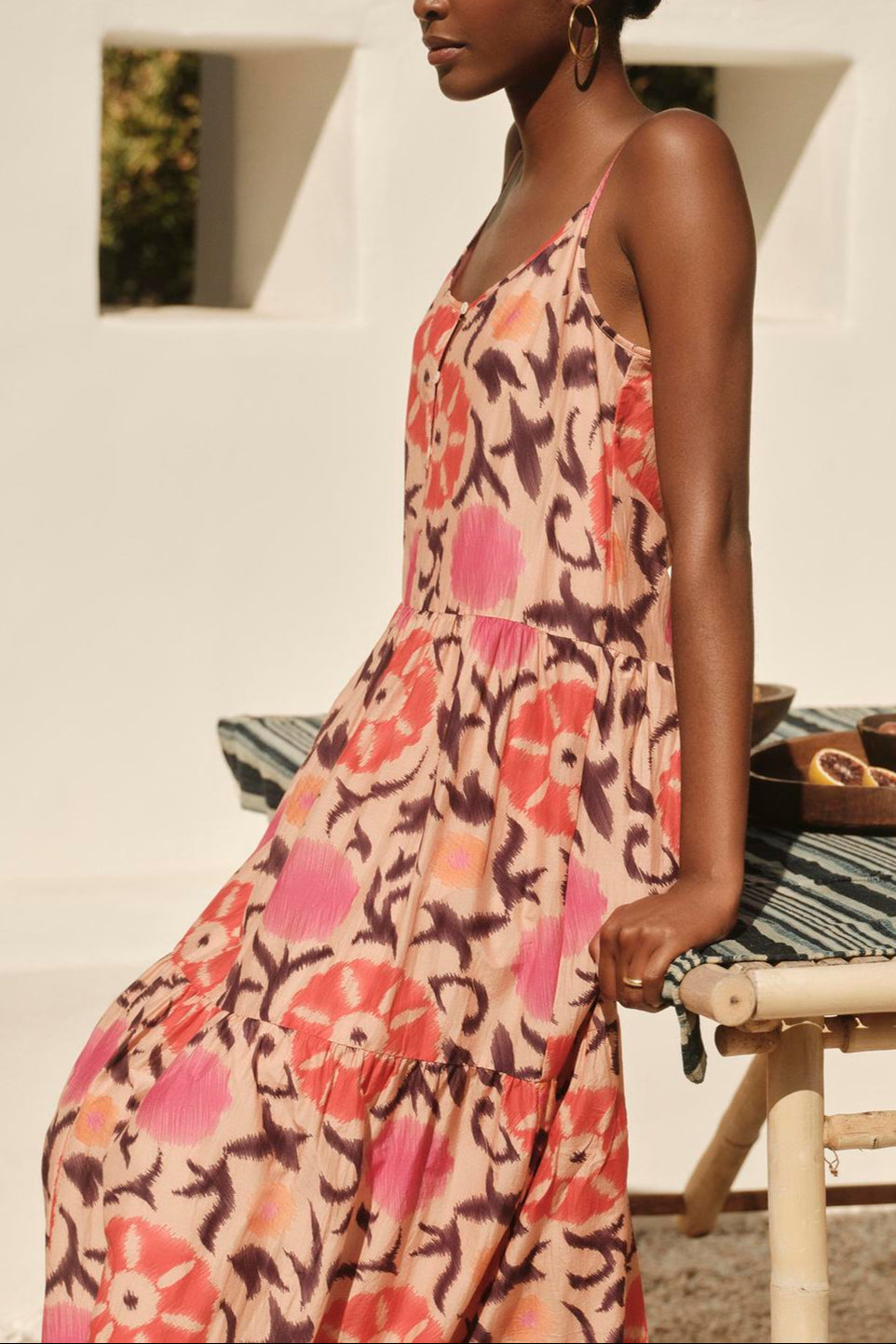 A woman in a KATE PRINTED SILK COTTON VOILE TANK DRESS by Velvet by Graham & Spencer stands beside a table with fruit, in a sunlit, white-walled courtyard.-36320311345345