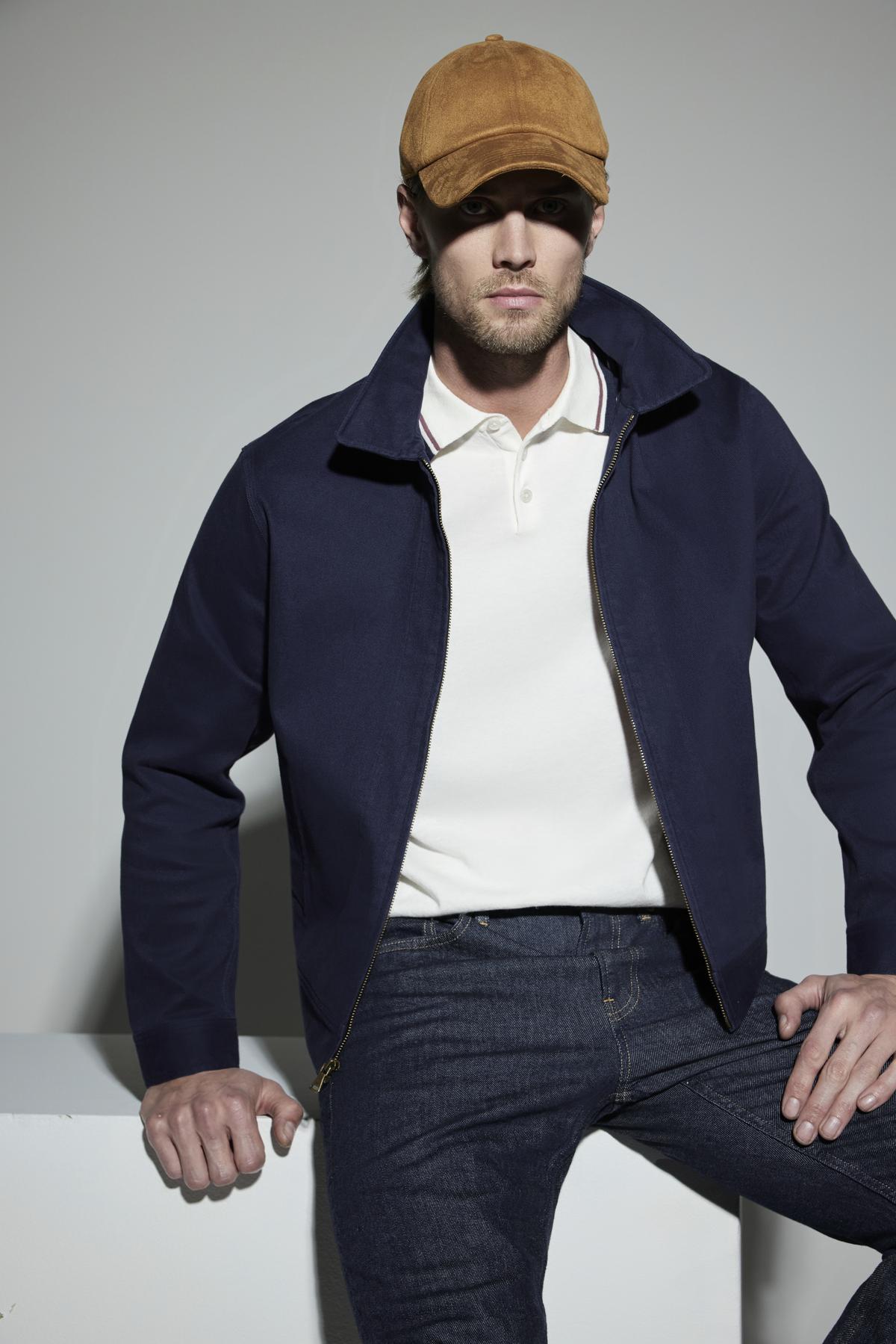 A man wearing a brown cap, white polo shirt, dark blue STEPHAN JACKET by Velvet by Graham & Spencer, and dark blue jeans is sitting on a white surface in a neutral setting.-37469116170433