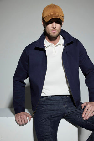 A man wearing a brown cap, white polo shirt, dark blue STEPHAN JACKET by Velvet by Graham & Spencer, and dark blue jeans is sitting on a white surface in a neutral setting.