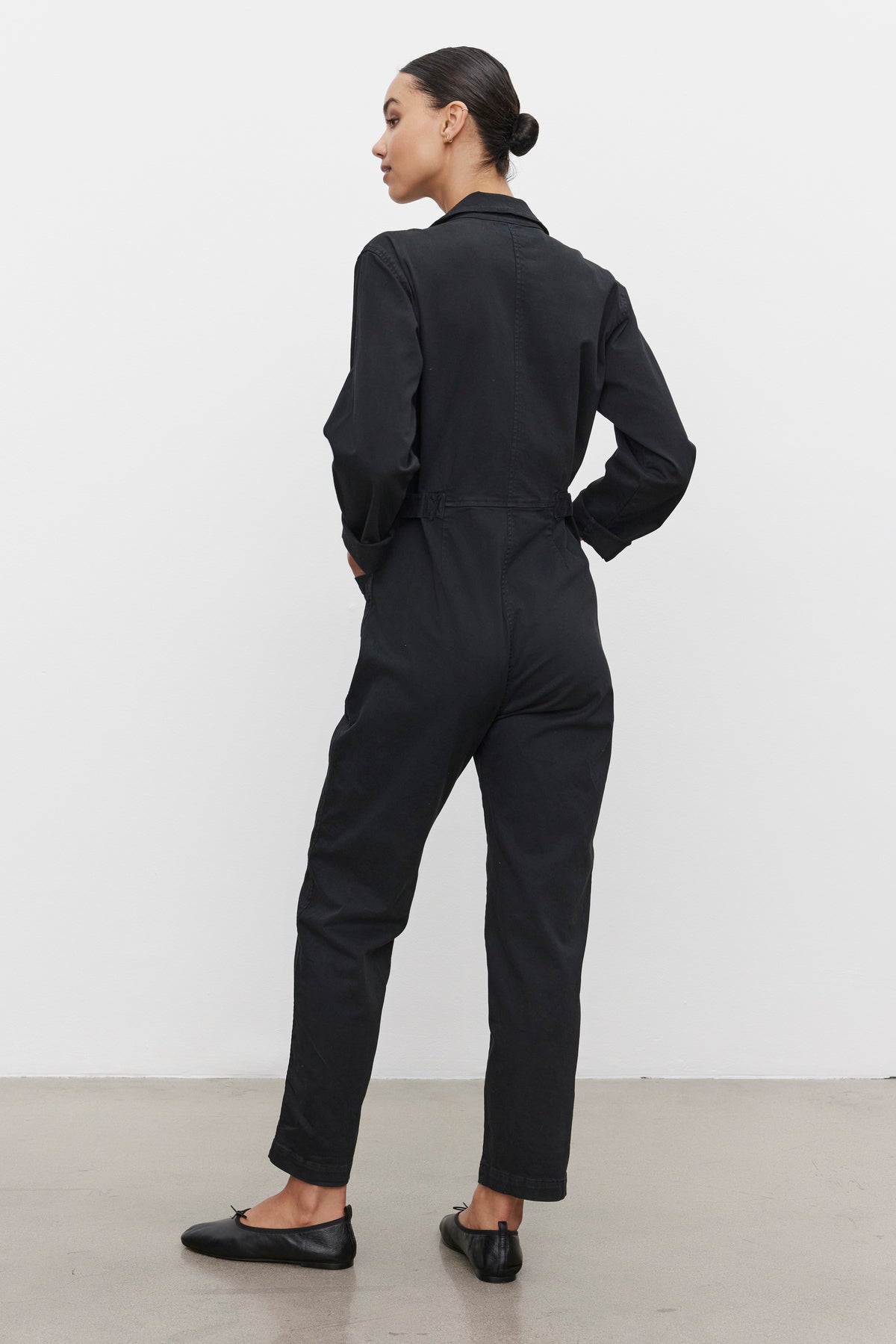 A person stands facing away, wearing the BRINLEY JUMPSUIT in black cotton twill by Velvet by Graham & Spencer and black flats, with their hair tied back, standing on a concrete floor against a plain white wall.