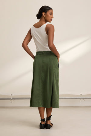 A person facing away, donning a white sleeveless top, an olive green LILIAN SKIRT by Velvet by Graham & Spencer made from stretch cotton twill, and black sandals stands in a neutral-toned room.
