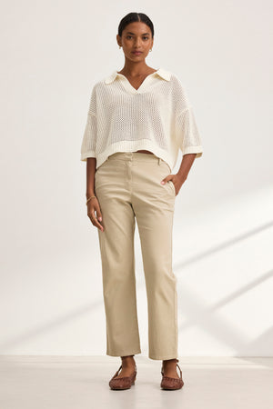 A person stands against a plain backdrop wearing a white knit sweater, Velvet by Graham & Spencer's beige MUSE PANT, and brown shoes.