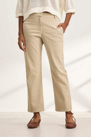 A person in a white sweater, MUSE PANT stretch cotton twill beige cropped pants by Velvet by Graham & Spencer, and brown shoes stands with one hand in their pocket against a light background.
