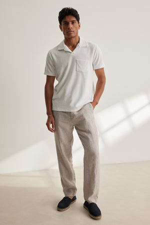 A man stands indoors against a plain light background wearing a white short-sleeve polo, Velvet by Graham & Spencer's JAE LINEN PANT with stripes and an elastic waistband, and dark slip-on shoes.