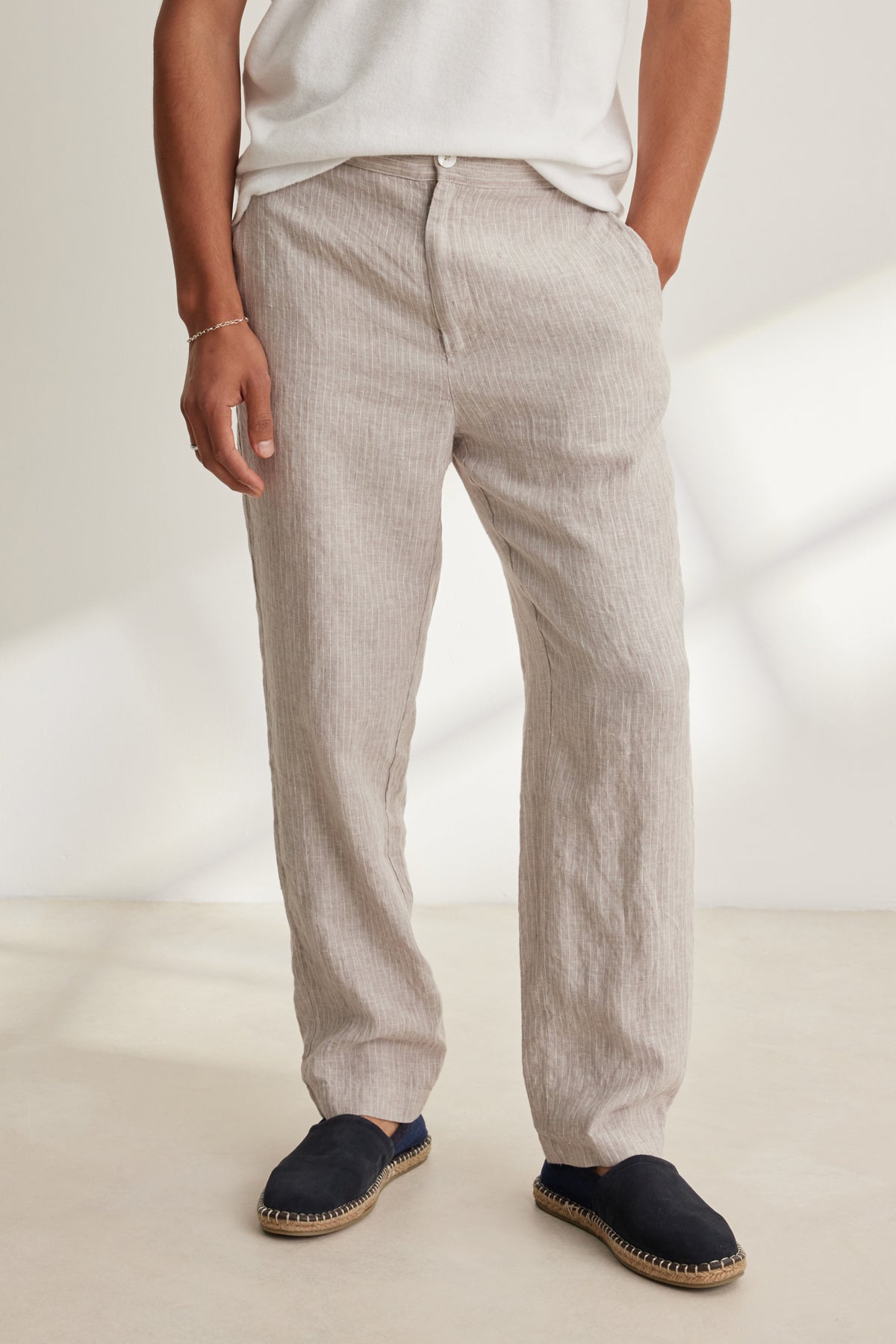 A person wears the Velvet by Graham & Spencer JAE LINEN PANT in light beige, featuring a rear elastic waistband, paired with a white shirt and black espadrilles, standing indoors with hands in pockets.-39152698196161