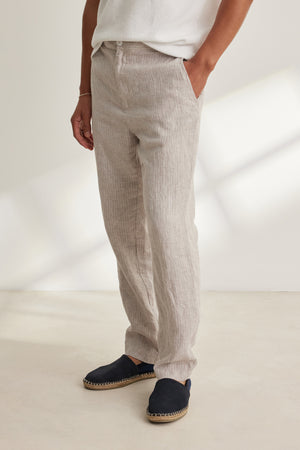 A person stands indoors wearing Velvet by Graham & Spencer’s JAE LINEN PANT in light beige with a rear elastic waistband, paired with a white shirt and black espadrilles, one hand in the pocket.