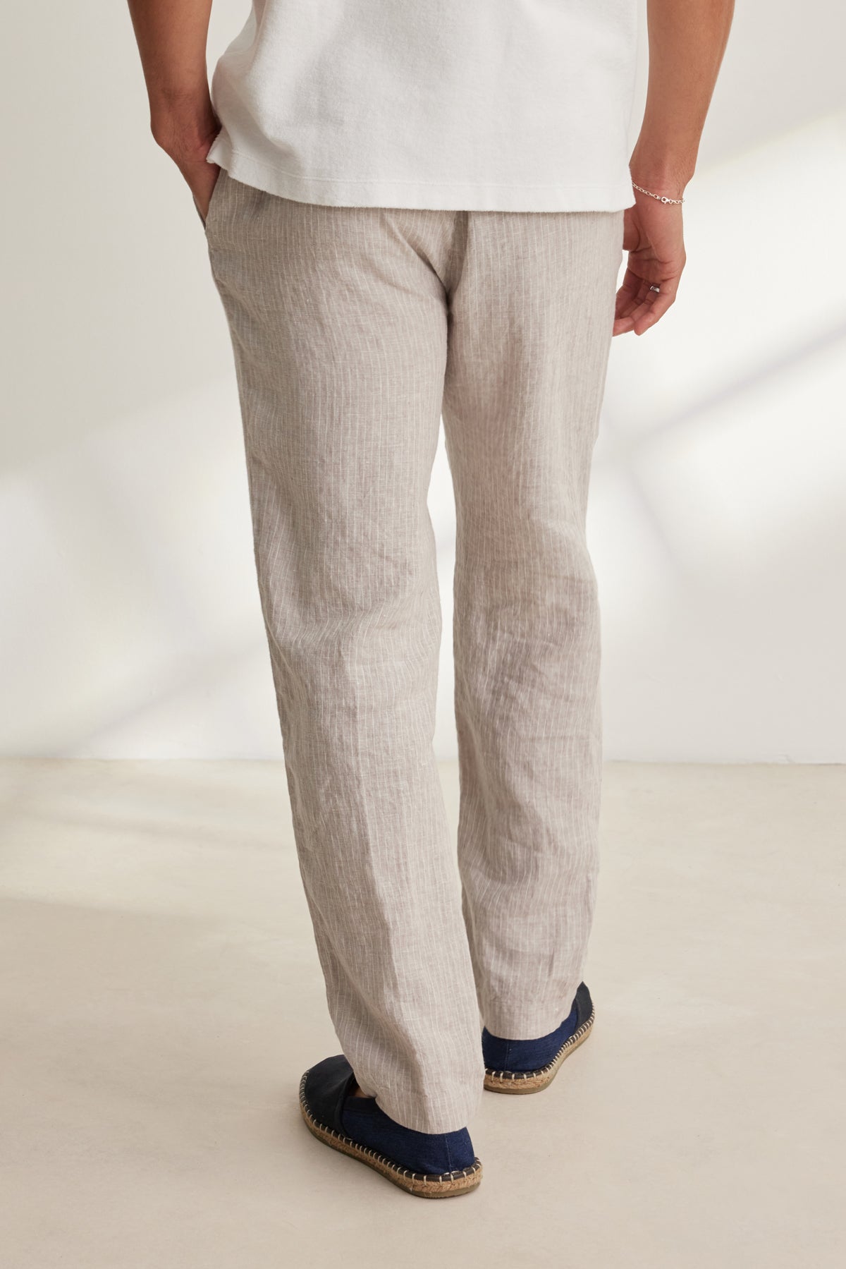 A person stands indoors with their back to the camera, wearing Velvet by Graham & Spencer's JAE LINEN PANT in light beige, featuring a straight-leg cut, paired with a white shirt and dark slip-on shoes.