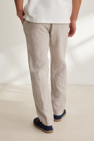 A person stands indoors with their back to the camera, wearing Velvet by Graham & Spencer's JAE LINEN PANT in light beige, featuring a straight-leg cut, paired with a white shirt and dark slip-on shoes.