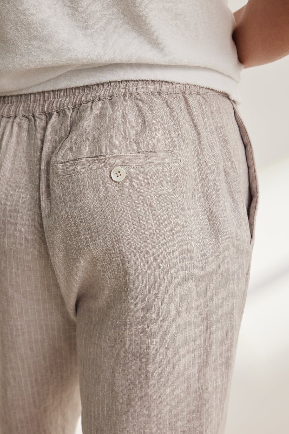 Close-up of a person wearing the JAE LINEN PANT from Velvet by Graham & Spencer—light beige, striped pants with a rear elastic waistband, buttoned back pocket, and straight-leg cut, paired with a white top for an effortless look.