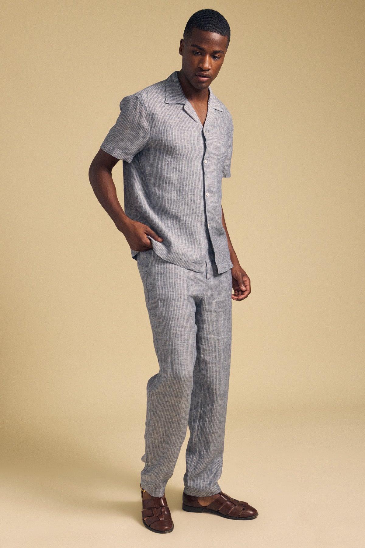 A man stands against a beige background wearing a short-sleeve, light grey striped shirt, JAE LINEN PANT by Velvet by Graham & Spencer—straight-leg, striped linen pants with an elastic waistband—and brown woven sandals.-41548474679489