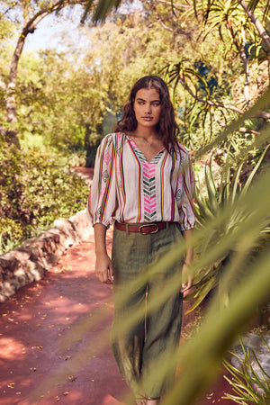 Woman walking down garden path wearing Beth Boho Top in multi colored jacquard print tucked into Dru pant in basil green with brown belt and necklace