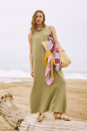 A woman in a axe green EDITH SLEEVELESS MAXI DRESS by Velvet by Graham & Spencer standing on long on the beach with a wicker basket.