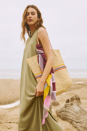 A woman wearing an IRIS TOTE by Velvet by Graham & Spencer, a lightweight green dress, and a colorful scarf walking on the beach.