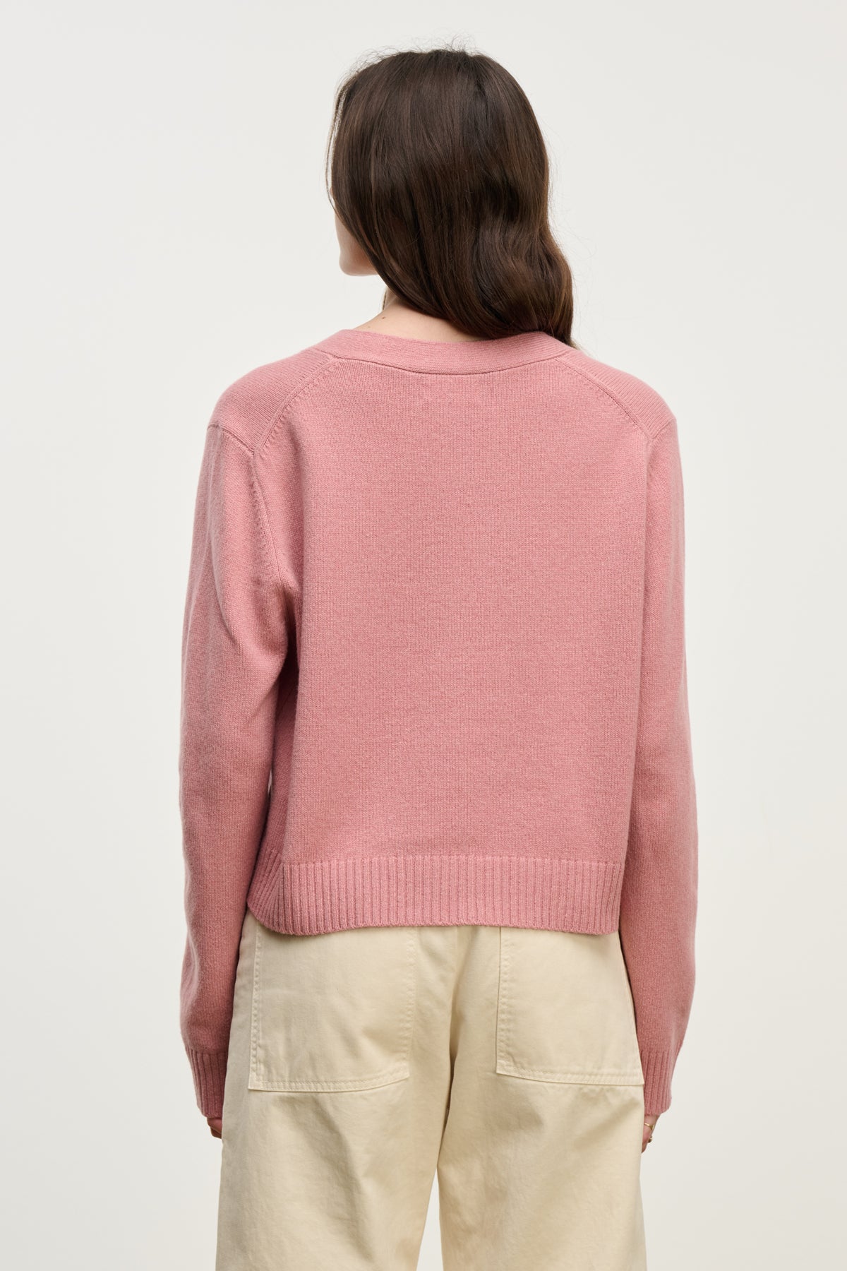 A person with long brown hair models the ETNA CARDIGAN by Velvet by Graham & Spencer in pink, paired with beige pants and shown from the back against a light background—an ideal cozy layer for every season.-40227054813377