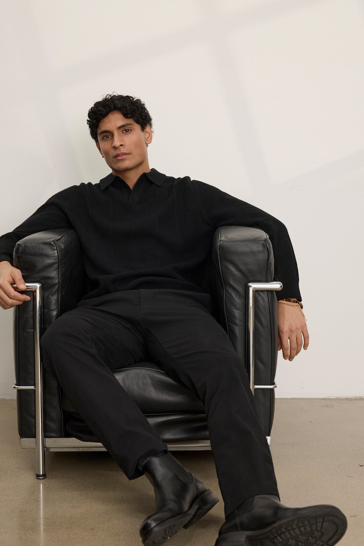 A person in the REDDING SWEATER POLO by Velvet by Graham & Spencer, black pants, and boots sits relaxed in a modern black leather armchair against a plain light wall, highlighting versatile layering in a minimalist setting.-40054247784641