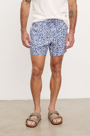 A man standing against a white background wearing a white t-shirt, Velvet by Graham & Spencer RICARDO SWIM SHORTS, and brown sandals. He has tattoos on his arms and is looking to the side.
