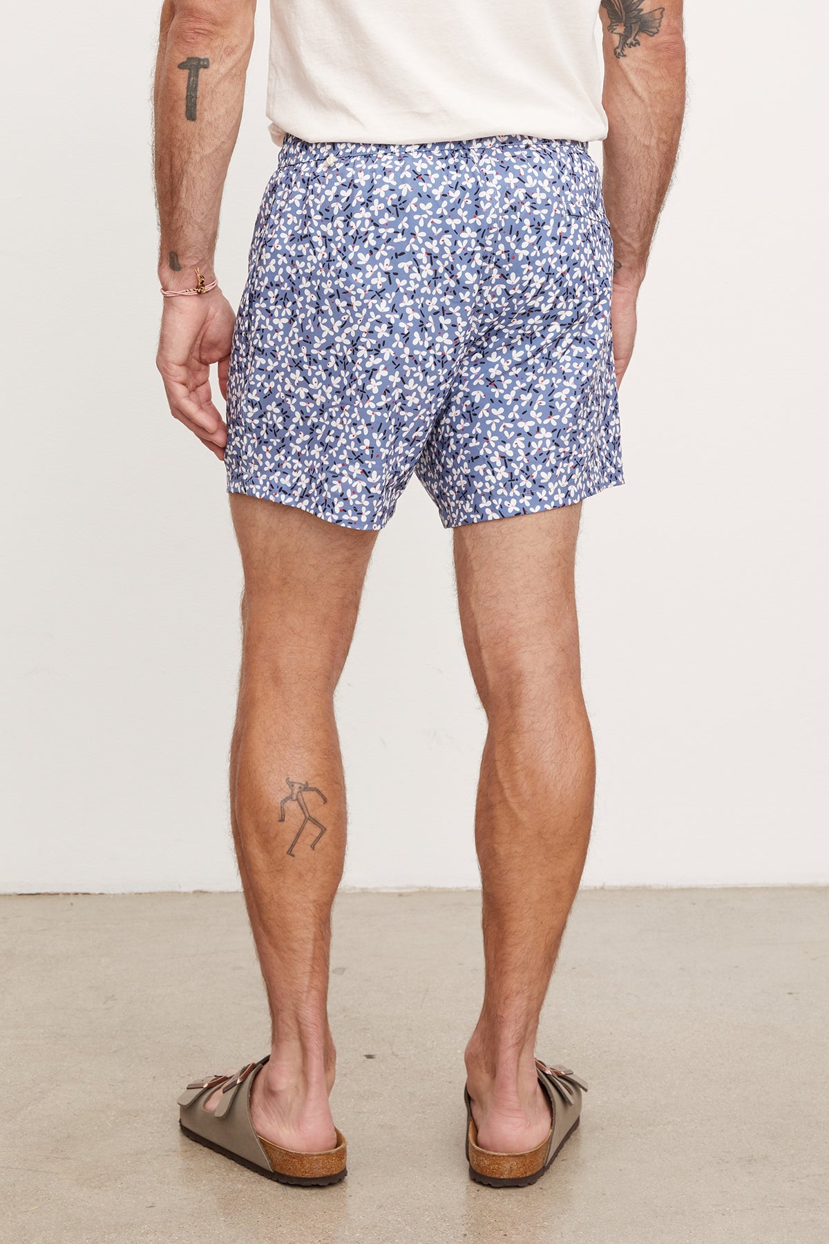 Man in Velvet by Graham & Spencer's RICARDO SWIM SHORT and brown flip-flops, standing with his back to the camera, displaying a tattoo on his right calf.