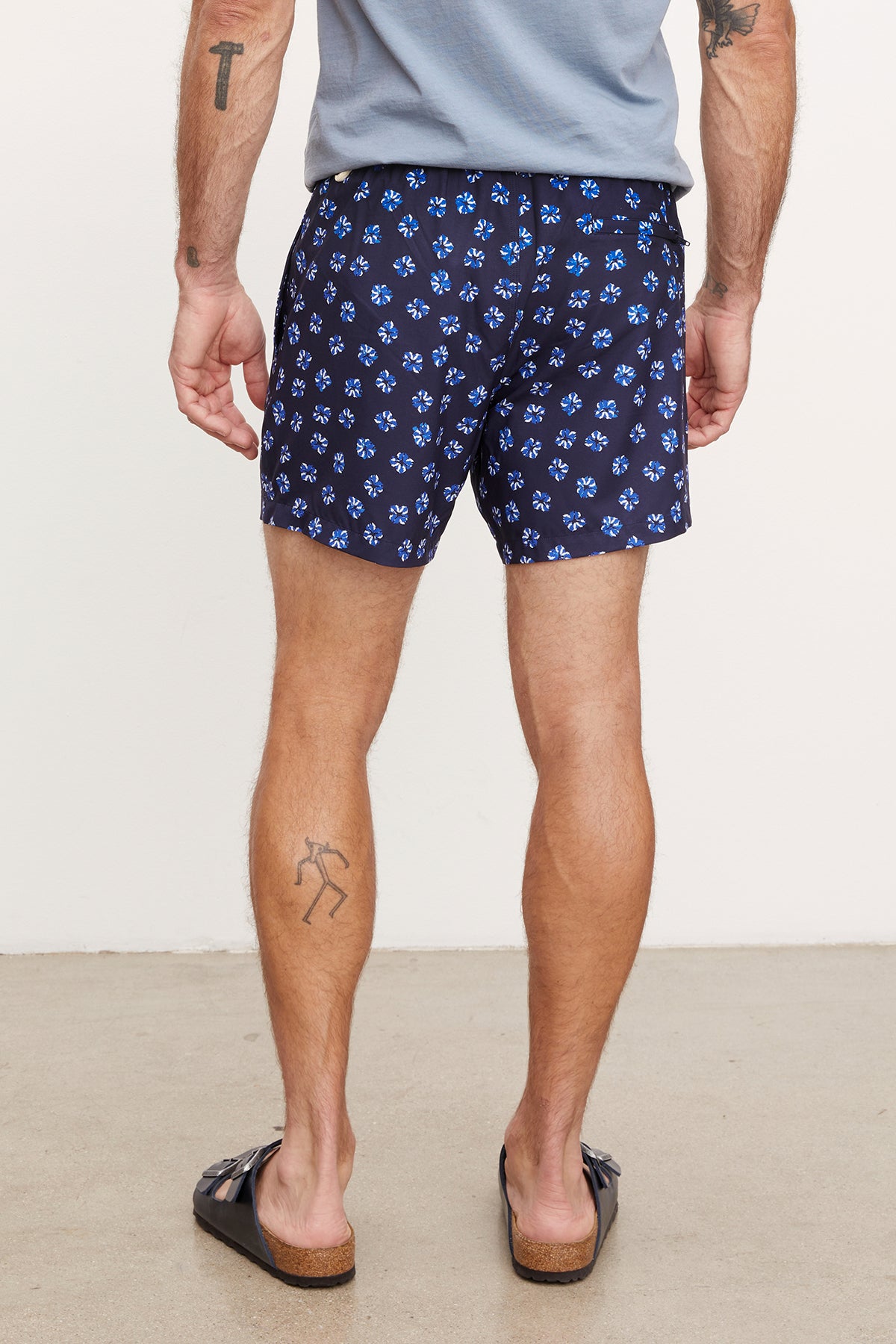 Man wearing Velvet by Graham & Spencer RICARDO SWIM SHORT swim shorts and black flip-flops, standing and showing the side and rear view, with visible tattoos on both arms and left leg.