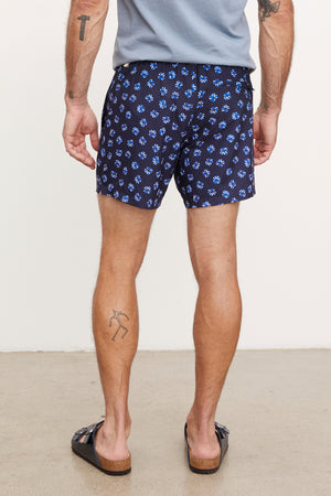 Man wearing Velvet by Graham & Spencer RICARDO SWIM SHORT swim shorts and black flip-flops, standing and showing the side and rear view, with visible tattoos on both arms and left leg.