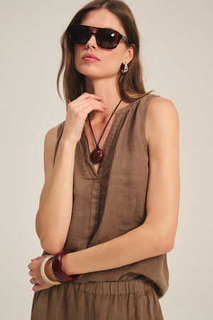 A woman wearing sunglasses and the TACY LINEN TANK TOP by Velvet by Graham & Spencer, featuring a brown v-neck sleeveless design and relaxed fit, paired with bold jewelry, stands against a neutral background.