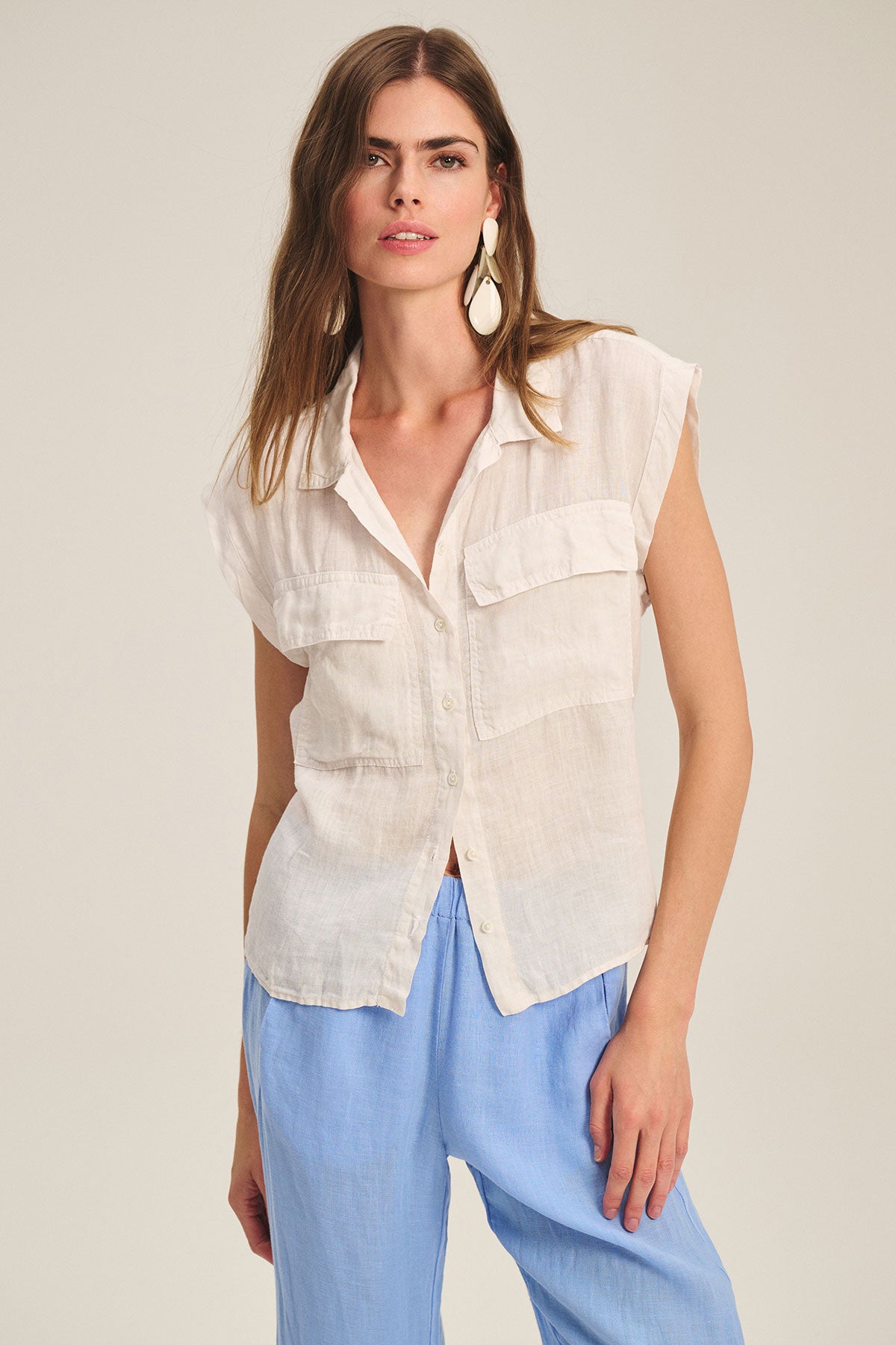 A woman wears the TASHA LINEN TOP by Velvet by Graham & Spencer, a sleeveless lightweight cargo style with flap pockets and buttons, paired with light blue pants against a plain background.-39725087654081