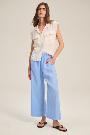 A woman stands against a plain background wearing the Velvet by Graham & Spencer LOLA LINEN PANT in light blue with an elastic waist, paired with a sleeveless white button-up shirt and black sandals.