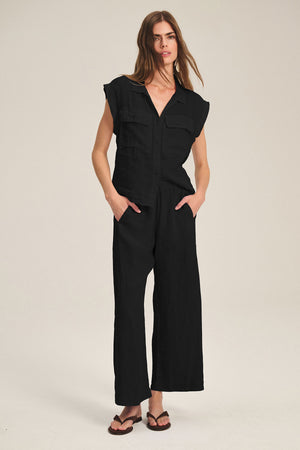 A woman stands with hands in pockets, wearing Velvet by Graham & Spencer's LOLA LINEN PANT—black pull-on linen trousers with an elastic waist—paired with a matching black short-sleeved button-up shirt and black flip-flops.