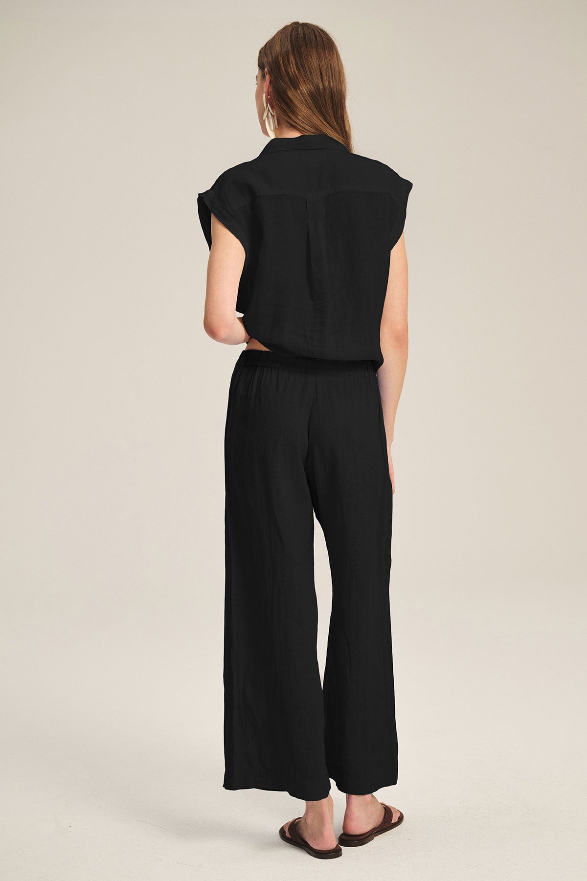 A woman faces away, wearing the TASHA LINEN TOP by Velvet by Graham & Spencer with matching black wide-leg pants and dark sandals against a plain light background.-39725082968257