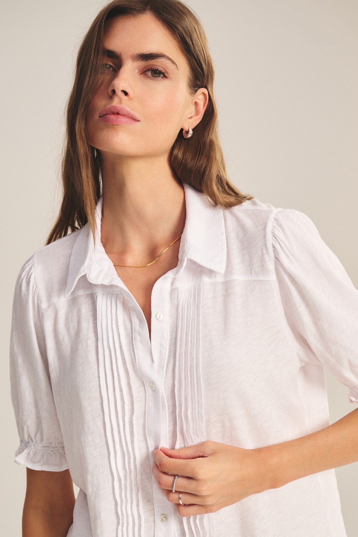 A woman wears the TATUM TEE by Velvet by Graham & Spencer—a white, button-front blouse with pleated details and subtle puff sleeves—standing against a neutral background.-41008418422977