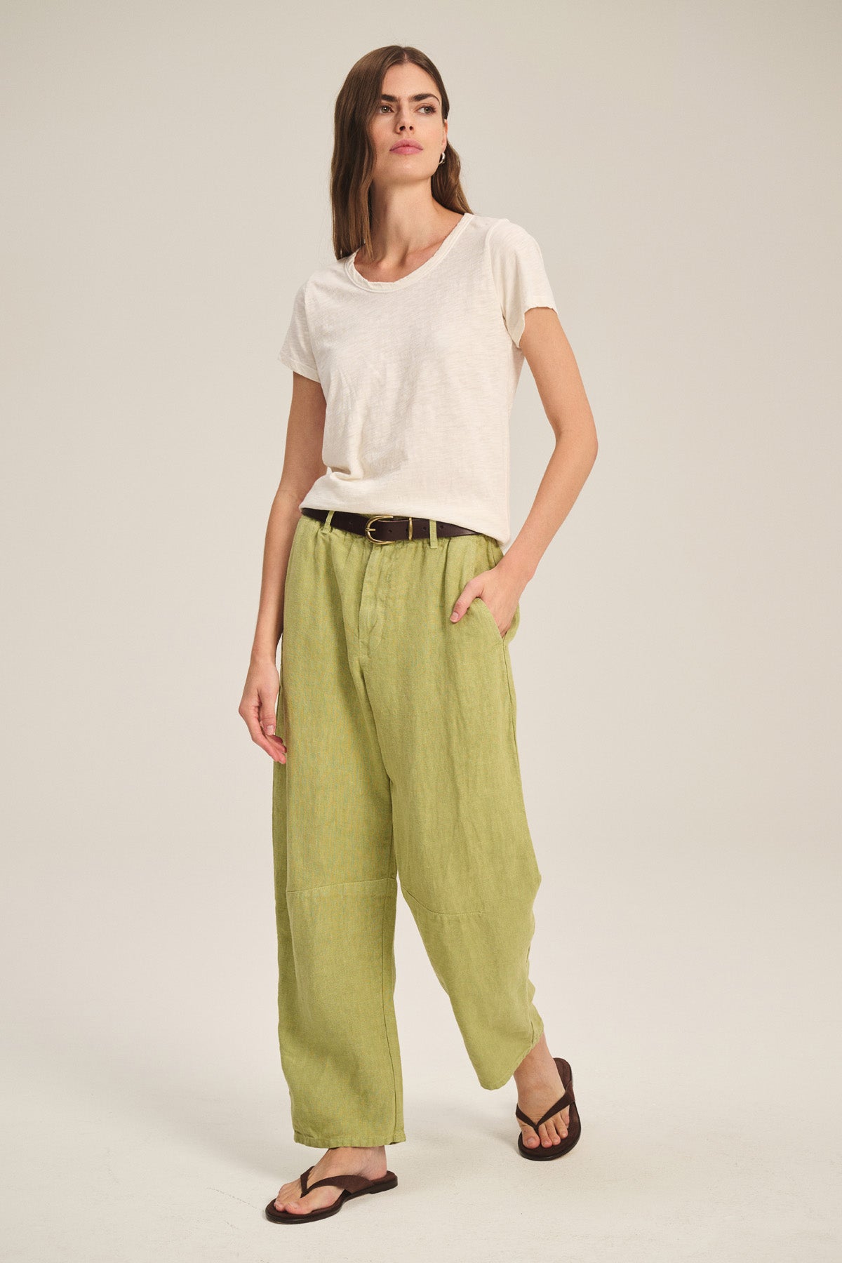 Person stands in a neutral studio setting wearing a white t-shirt, Velvet by Graham & Spencer TAYLOR HEAVY LINEN PANT in light green, a dark belt, and brown sandals.-41436030402753