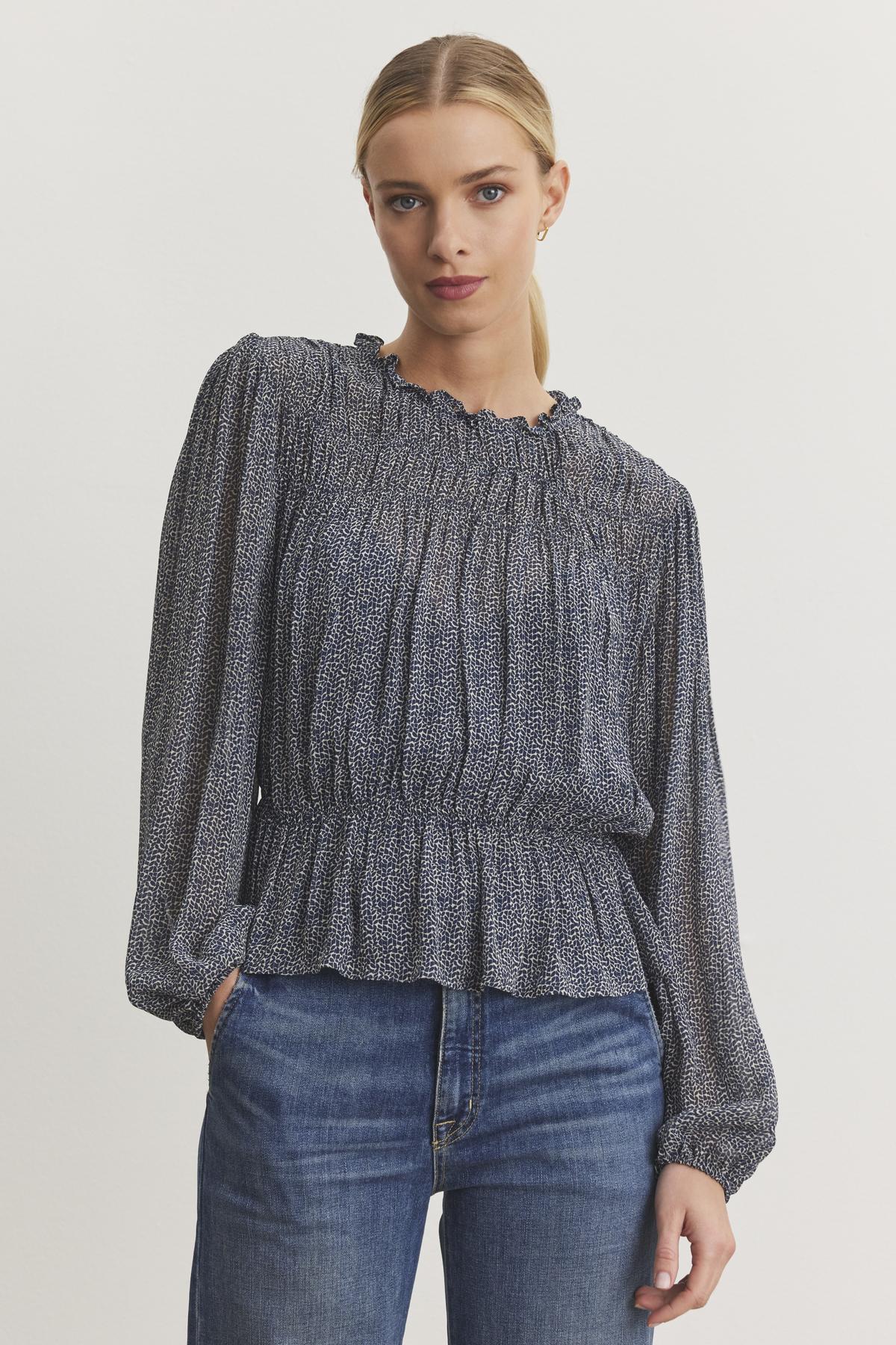 A person wearing the BLEN TOP from Velvet by Graham & Spencer, featuring a long-sleeve design with a smocked elastic waist and ruffle neckline, paired with blue jeans, stands against a plain background.-38246328303809