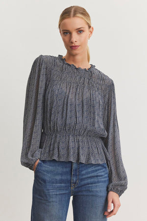 A person wearing the BLEN TOP from Velvet by Graham & Spencer, featuring a long-sleeve design with a smocked elastic waist and ruffle neckline, paired with blue jeans, stands against a plain background.