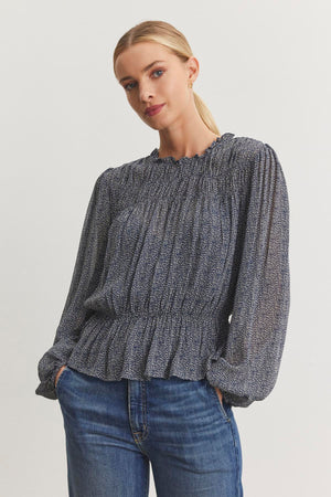 A woman wearing the BLEN TOP by Velvet by Graham & Spencer, featuring a ruffle neckline and a patterned design, paired with blue jeans, her hands casually resting in her pockets.