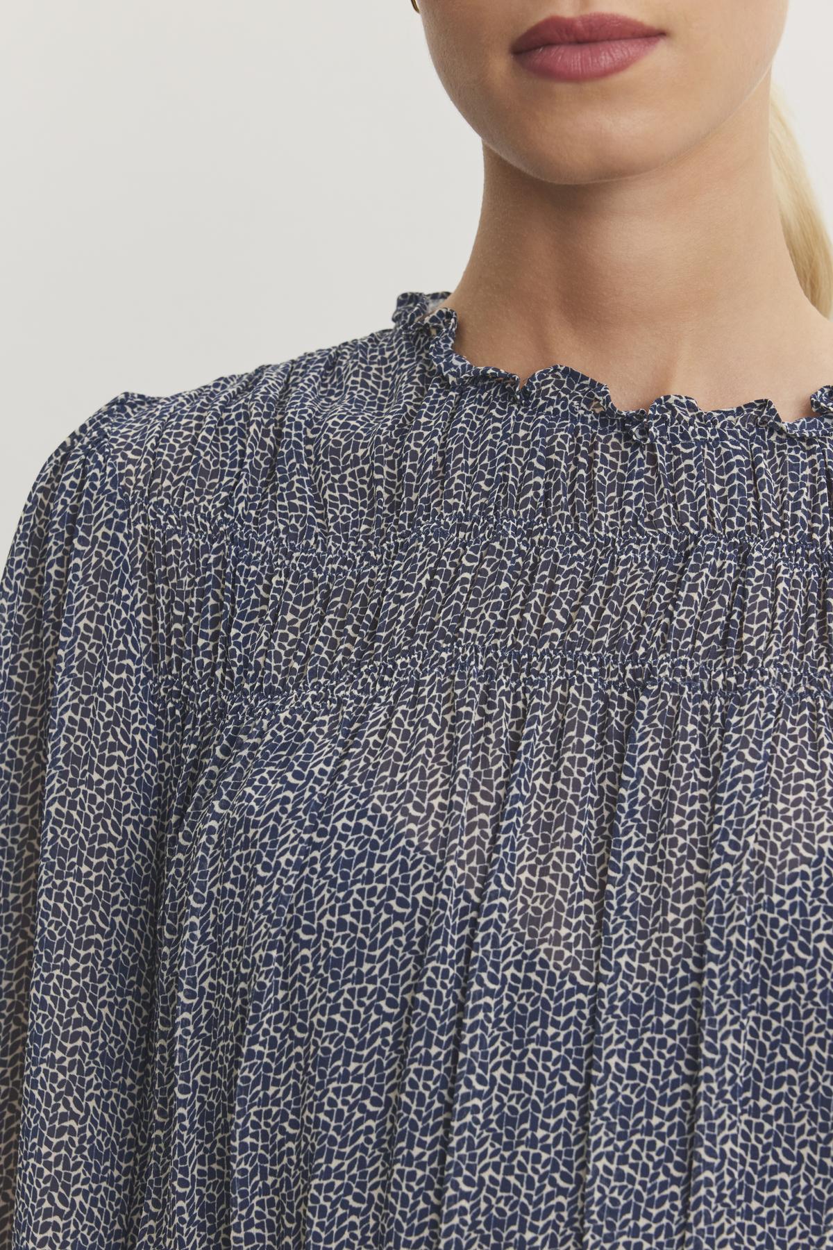 A close-up of a person wearing the Velvet by Graham & Spencer BLEN TOP, featuring a blue and white pattern with a smocked elastic and ruffled neckline.-38246328434881