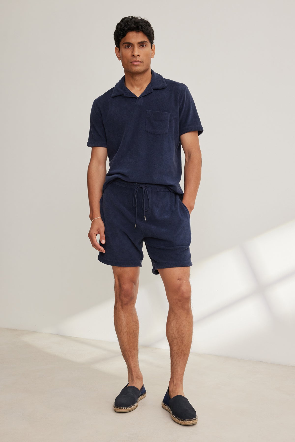 A man stands indoors wearing Velvet by Graham & Spencer’s SALEM SHORT—navy cotton terry cloth lounging shorts with a drawstring waist—paired with a matching short-sleeve shirt and navy slip-on shoes.-39152734306497
