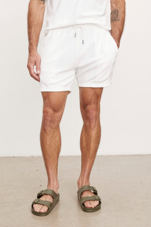 A man in a white t-shirt stands on a beige floor, his hands in the pockets of Velvet by Graham & Spencer's SALEM SHORT, featuring a drawstring elastic waist. He complements his look with green sandals.