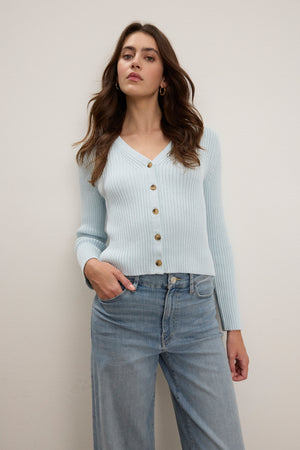 A person wearing the HYDIE CARDIGAN by Velvet by Graham & Spencer, a light blue ribbed knit with button closure, and wide-leg jeans stands against a plain backdrop.