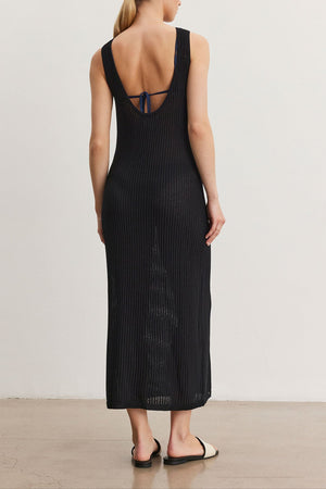 A woman stands facing away, wearing a CALLEN DRESS by Velvet by Graham & Spencer, black and ribbed with a low back and small bow detail. She has blonde hair tied in a low bun and wears black flat sandals.
