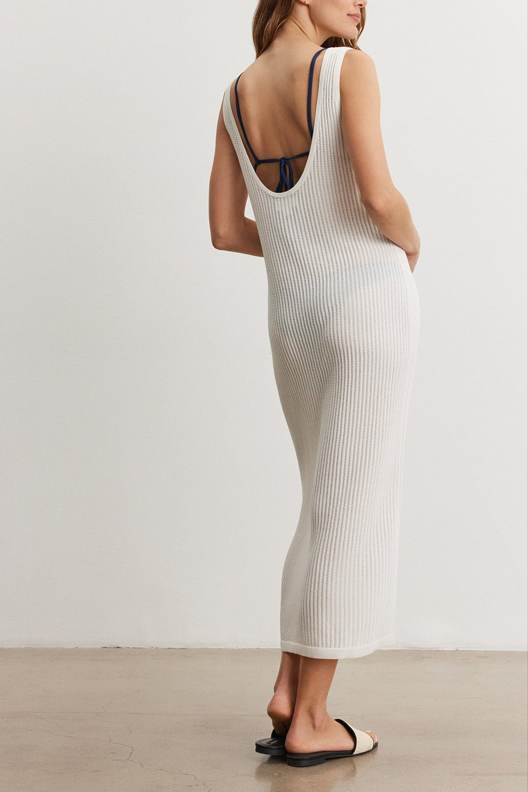 A woman in a Velvet by Graham & Spencer CALLEN DRESS stands facing away, glancing over her shoulder, in a sparse room with a concrete floor.