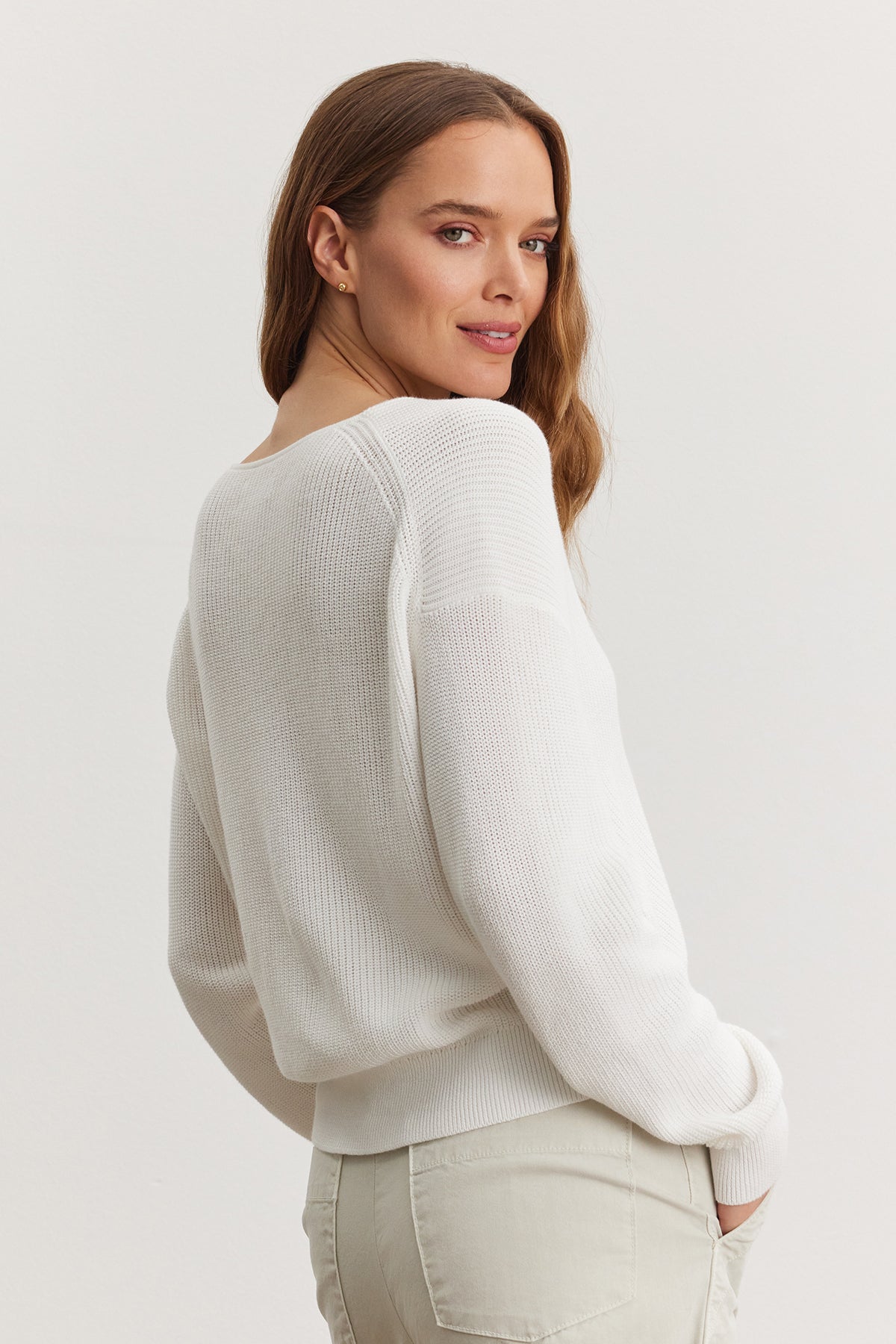 A woman in a white ribbed Velvet by Graham & Spencer TAVA cardigan and light beige pants, standing with her back slightly turned, looking over her shoulder with a soft smile.-36909564166337