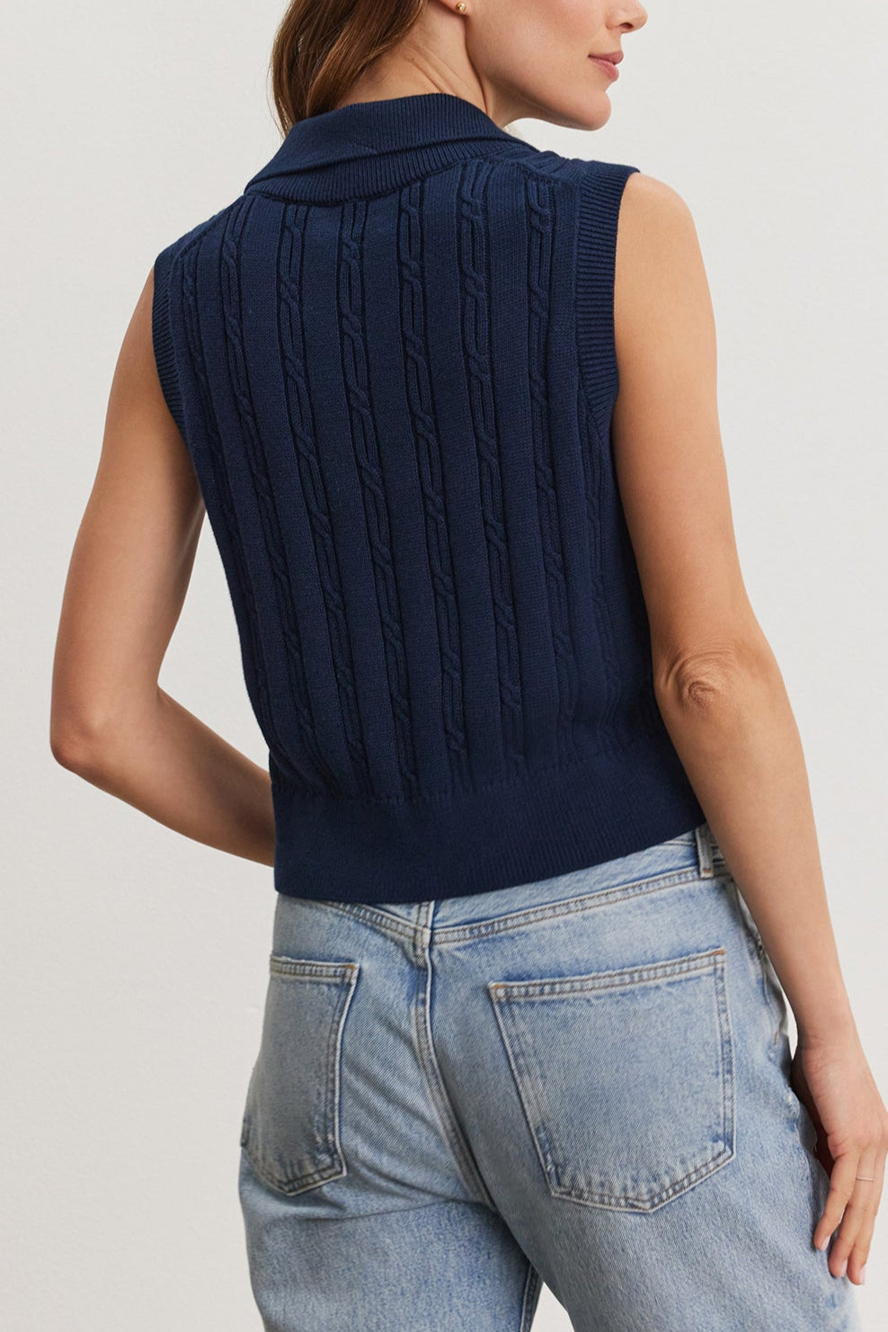 Woman standing facing away from the camera, wearing a blue Velvet by Graham & Spencer Wendy Sweater Vest and light blue jeans, focusing on the sweater's cable knit design.-36909568524481