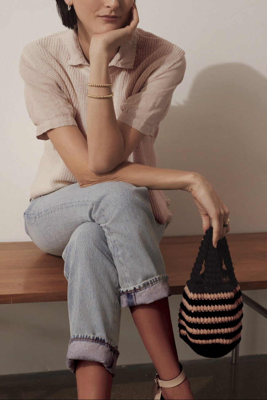 A woman sits on a wooden bench, resting her chin on her hand, wearing a pink blouse, jeans, and holding a Velvet by Jenny Graham KNOX BAG.-36890823262401