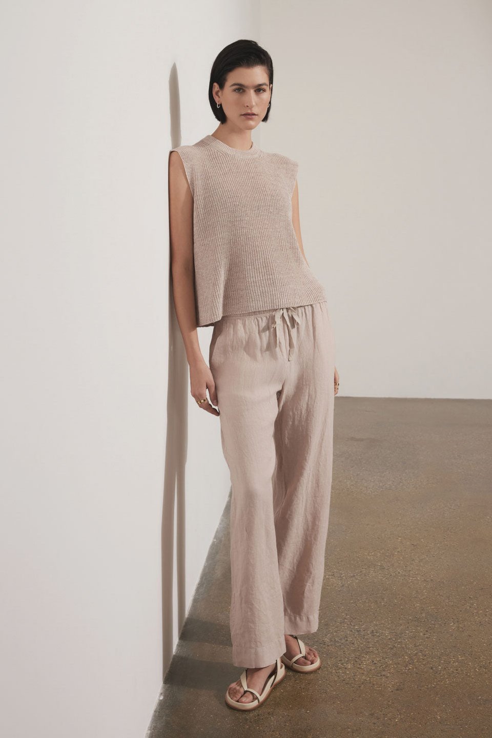 A woman standing against a white wall wearing a sleeveless beige mesh top and matching relaxed fit Velvet by Jenny Graham PICO LINEN PANT with sandals.-36863283462337