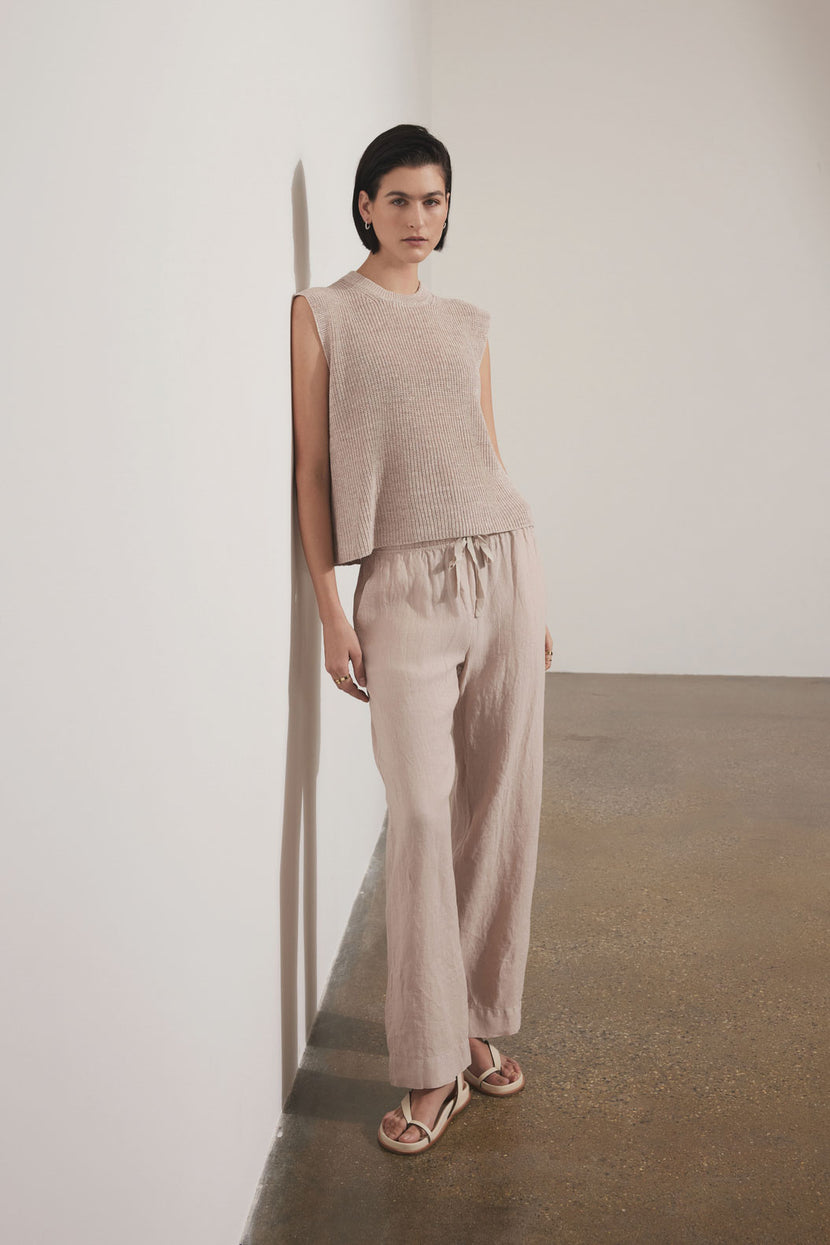 A woman standing against a white wall wearing a sleeveless beige mesh top and matching relaxed fit Velvet by Jenny Graham PICO LINEN PANT with sandals.