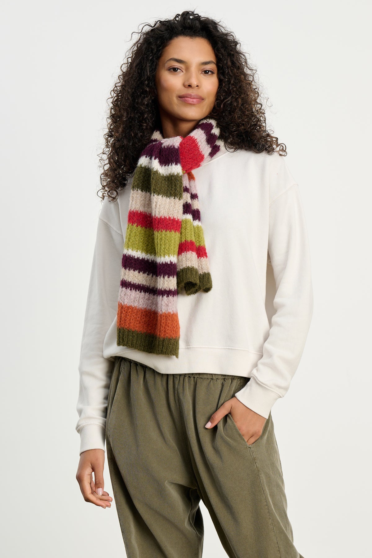 A person wearing a cream sweatshirt and olive pants accessorizes with the LUCA ALPACA SCARF by Velvet by Graham & Spencer, adding a colorful, textured touch, while standing against a plain background.-39742257889473
