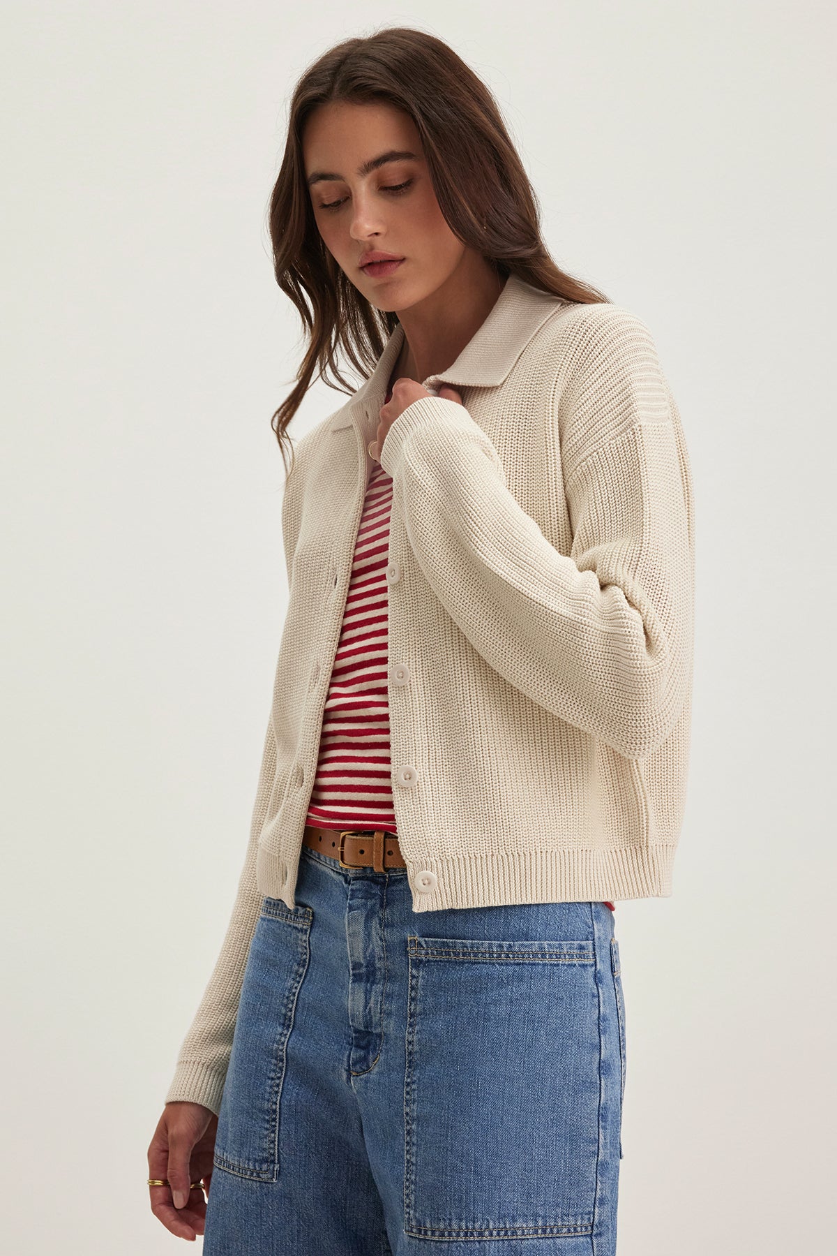 A woman wearing Velvet by Graham & Spencer's TAJ CARDIGAN in beige-textured cotton pairs it with a red and white striped shirt and blue jeans while standing with her eyes closed against a plain background.-38663164461249