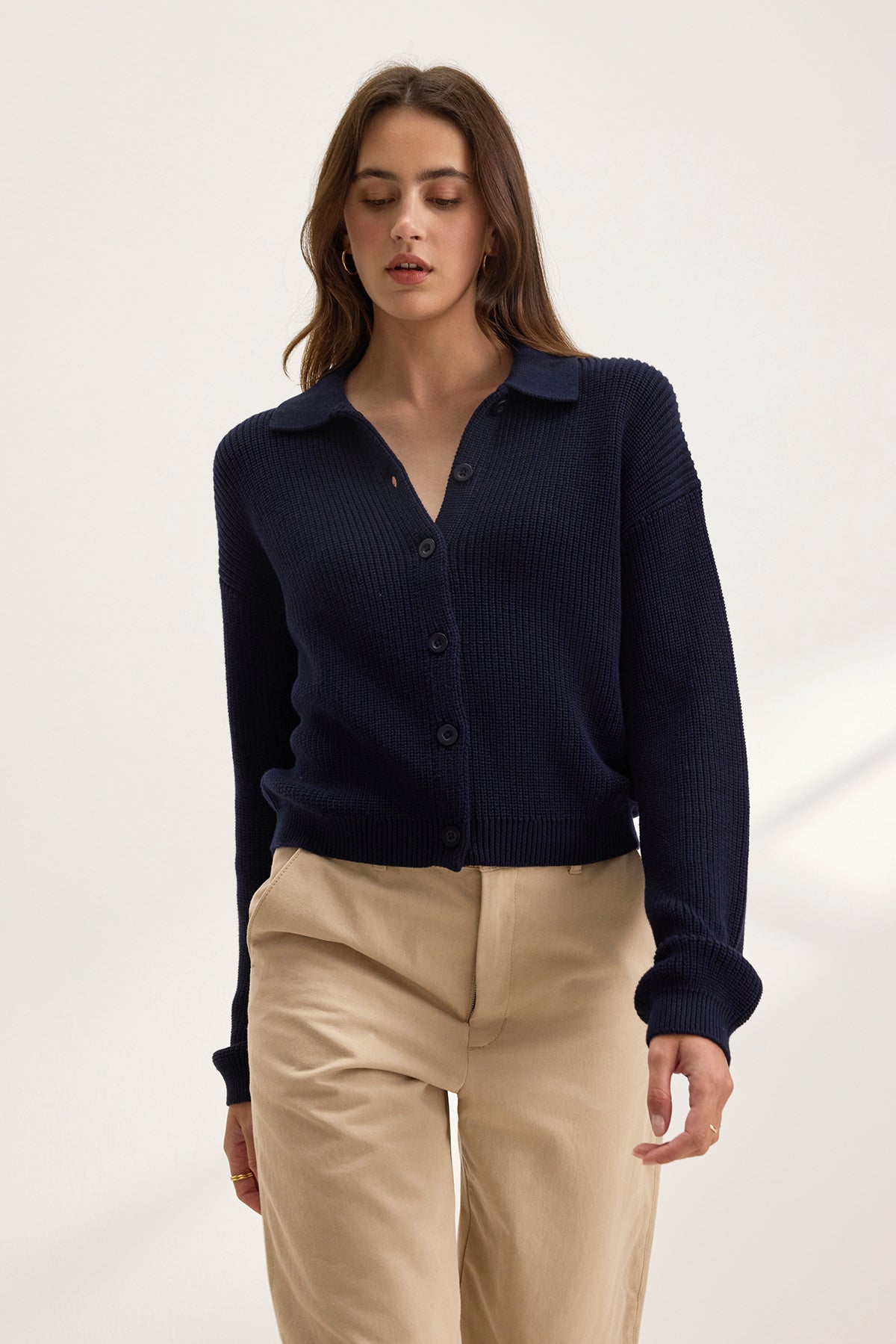 A person in a dark blue TAJ CARDIGAN by Velvet by Graham & Spencer, featuring textured cotton and cropped length, paired with beige pants, stands against a neutral background.