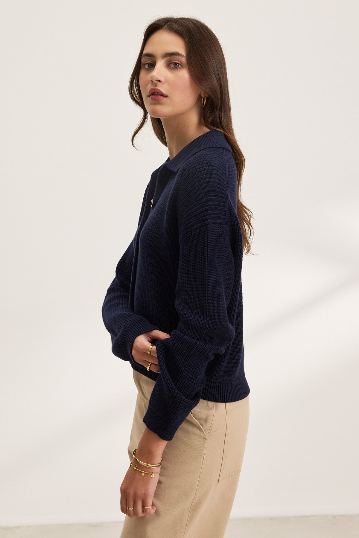 A person with long hair wears the TAJ CARDIGAN by Velvet by Graham & Spencer in a navy hue and beige pants. They're standing sideways against a plain background, looking at the camera to showcase their cropped outfit.-38663164133569
