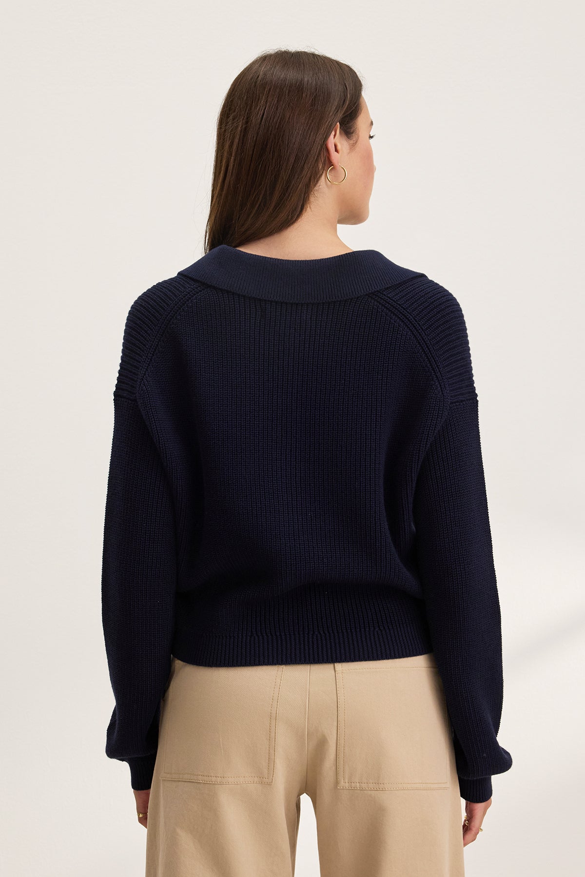 Rear view of a person with long brown hair wearing the cropped navy TAJ CARDIGAN by Velvet by Graham & Spencer, paired with beige pants, against a plain background.-38663164166337